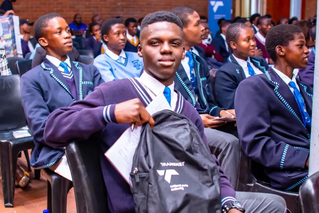 A student in an assembly hall, holding up a Transnet branded bag to have his picture taken