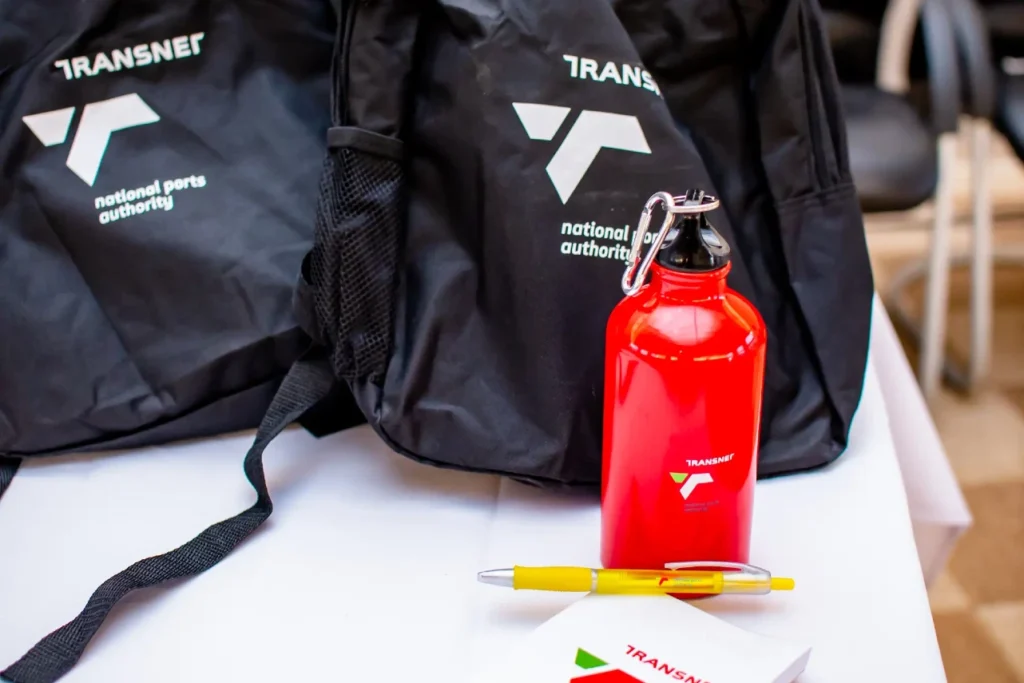 various transet branded apparel on a display table including 2 bags, a hot water bottle and branded pen and notpad